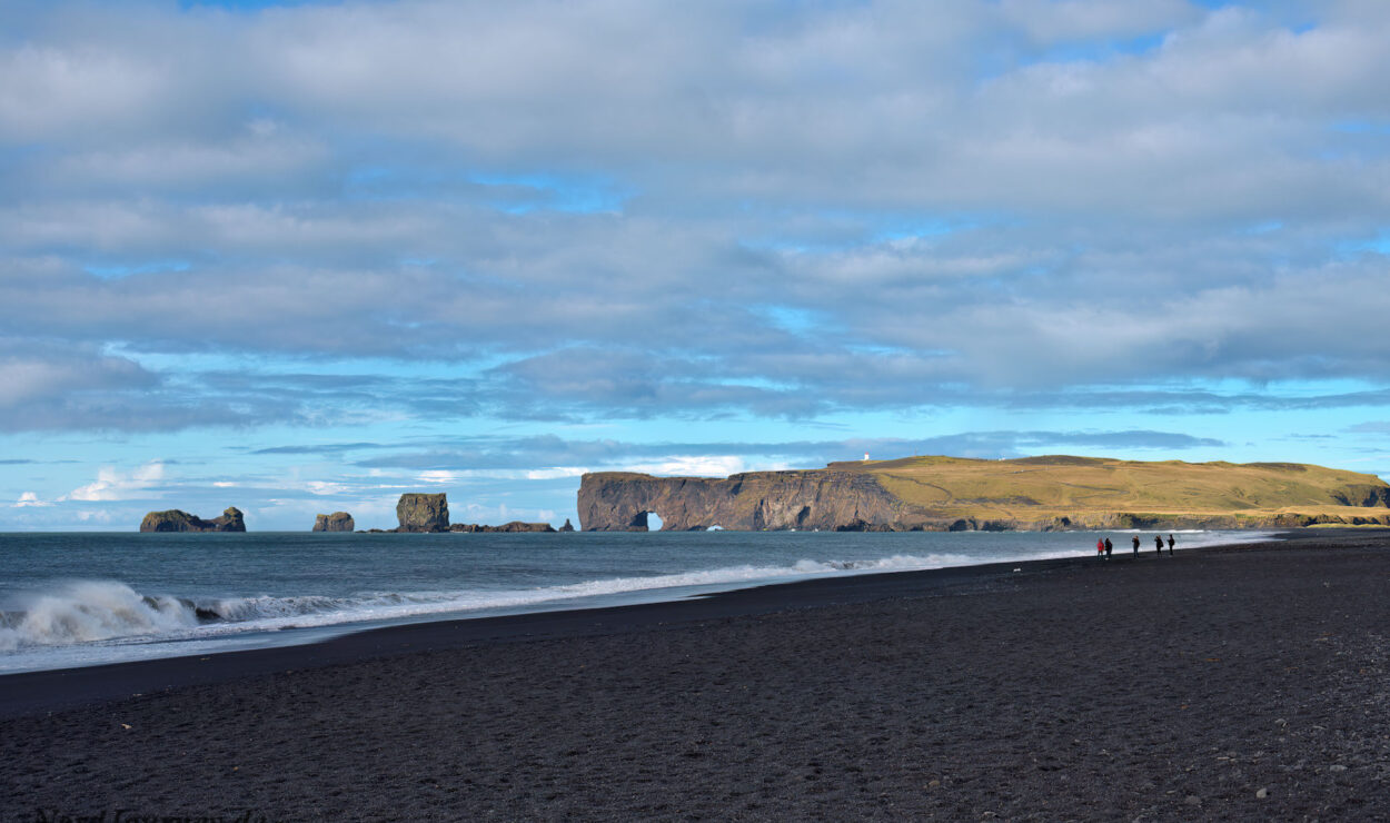 Schwarzer Sandstrand mit sanften Wellen, Felsformationen im Meer, eine grasbewachsene Klippe in der Ferne und ein paar Menschen, die unter einem bewölkten blauen Himmel am Ufer entlang spazieren.