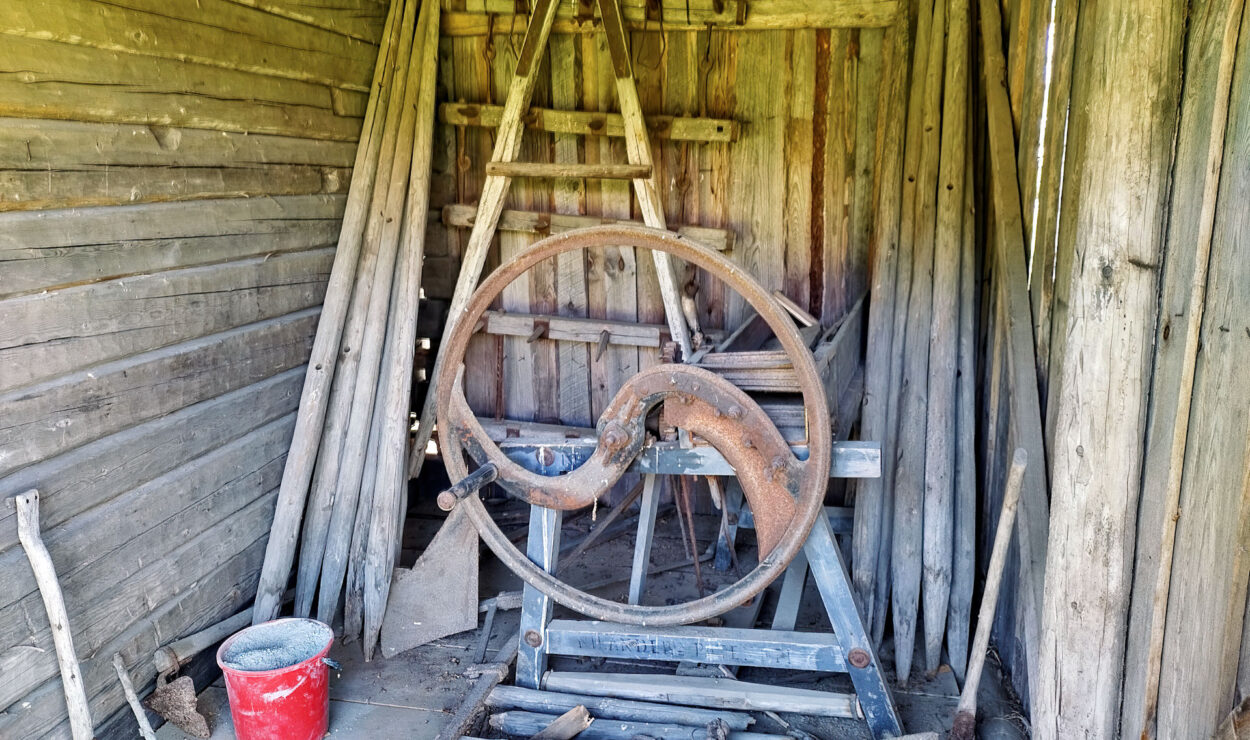 Rustikaler Holzschuppen-Innenraum mit verschiedenen Holzbrettern und einem alten, verrosteten Metallwerkzeug oder Rad auf einem Ständer. Auf dem Holzboden steht ein roter Eimer.