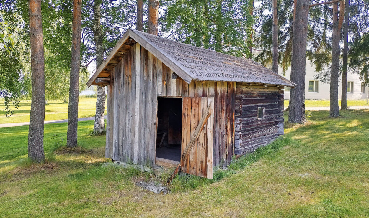 Ein kleiner, rustikaler Holzschuppen mit teilweise geöffneter Tür steht inmitten hoher Bäume auf einer Rasenfläche, im Hintergrund ist ein weißes Gebäude zu sehen.