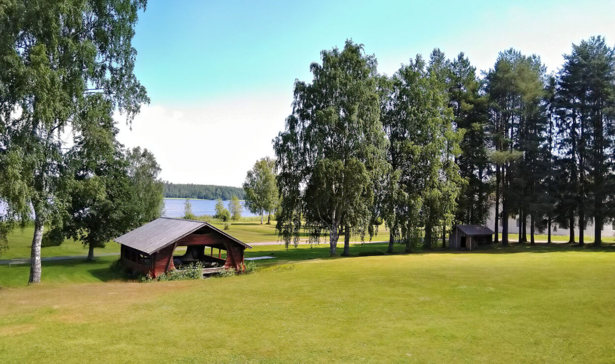 Eine Holzhütte steht auf einer von Bäumen umgebenen Rasenfläche. Im Hintergrund ist unter einem klaren Himmel ein See zu sehen.