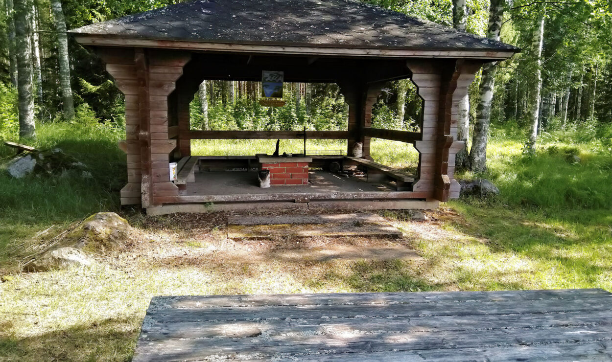 Holzpavillon mit Picknicktisch in einer Waldumgebung.