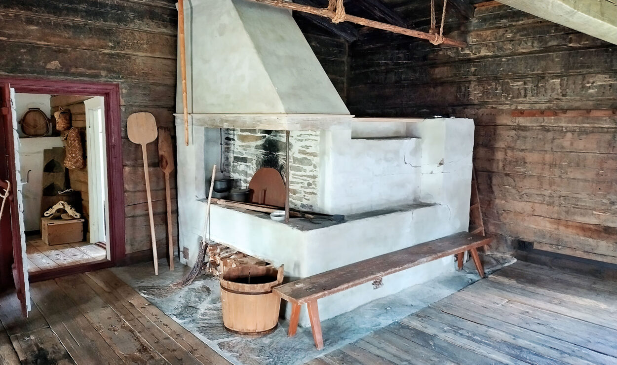 Eine rustikale, historische Küche mit einem großen Kamin aus Stein und Gips, einer Holzbank und einem Eimer auf einem Holzboden. Durch die Tür gelangt man in einen weiteren Raum mit Holzdekor.