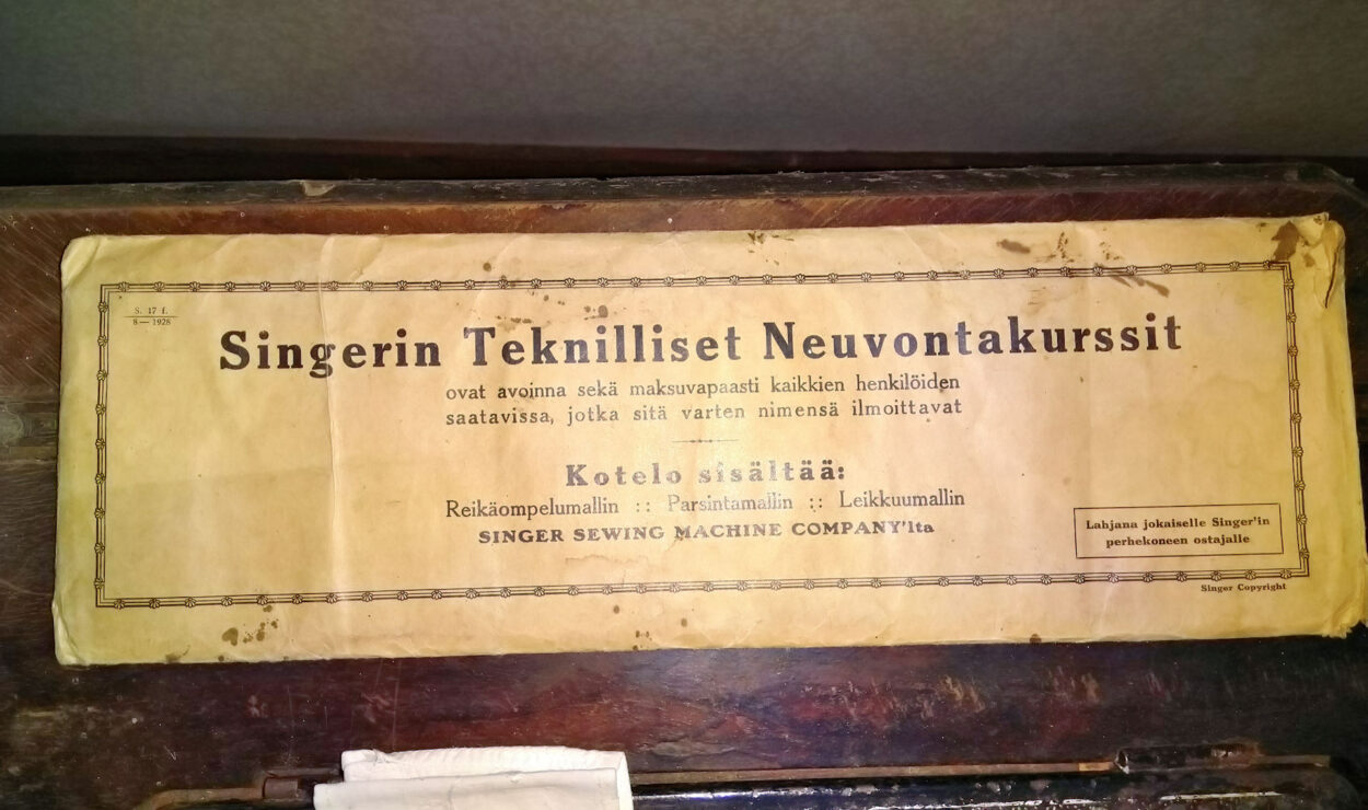 Eine alte Hülle für eine Singer-Nähmaschine mit einem Text auf Finnisch zu technischen Beratungskursen, in dem die Verfügbarkeit und Einzelheiten zu den Komponenten in der Schachtel erwähnt werden.