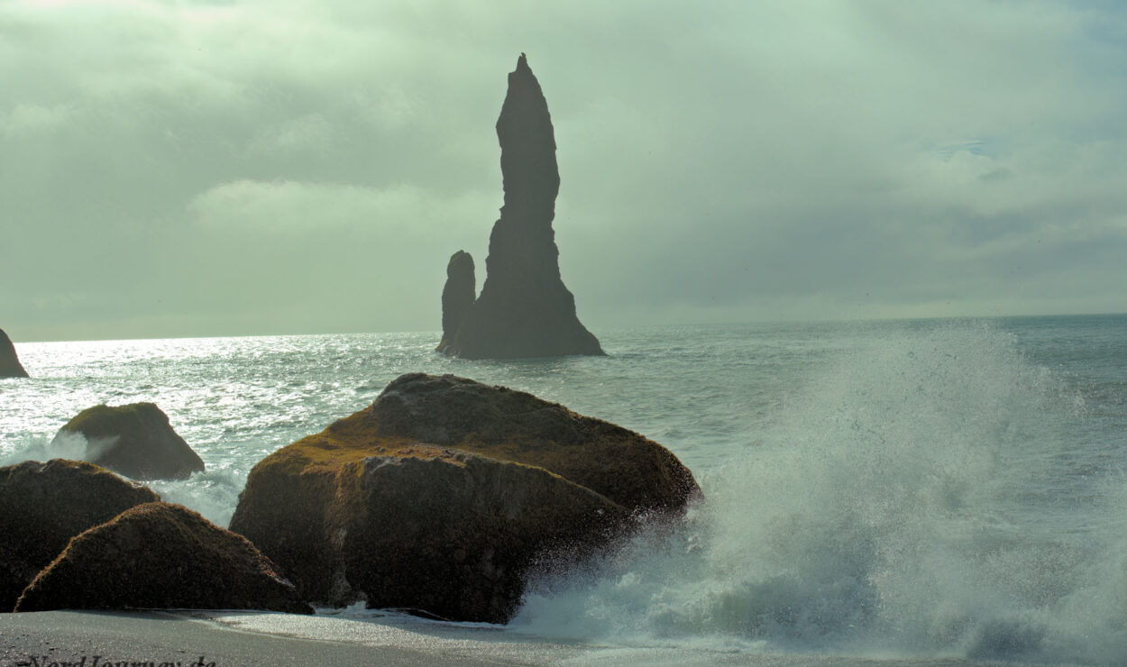 Wellen schlagen gegen Felsen an einem Strand, im Hintergrund ist unter einem bewölkten Himmel ein hoher, spitzer Felspfeiler zu sehen.