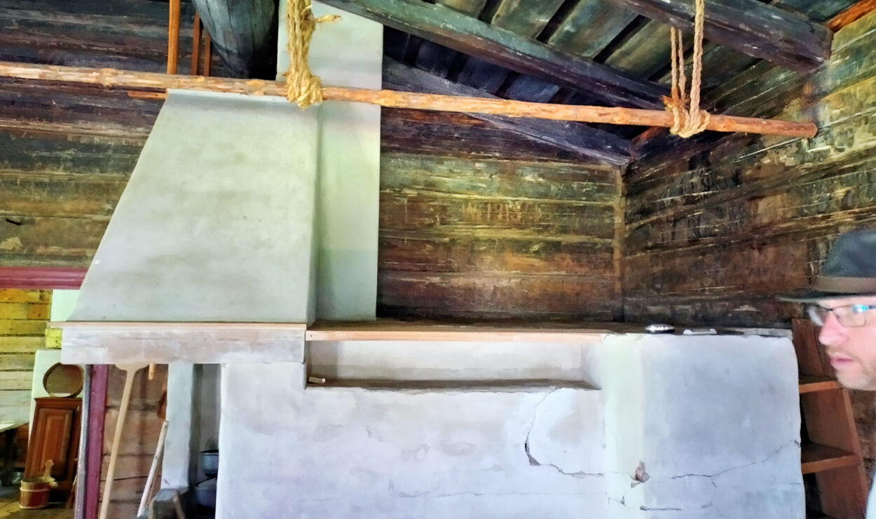 Rustikales Holzinterieur mit großem Steinkamin und an einem Seil aufgehängtem Holzbalken. Eine Holzdecke und Blockwände rahmen den Raum ein. Auf der rechten Seite ist teilweise eine Person mit Hut zu sehen.