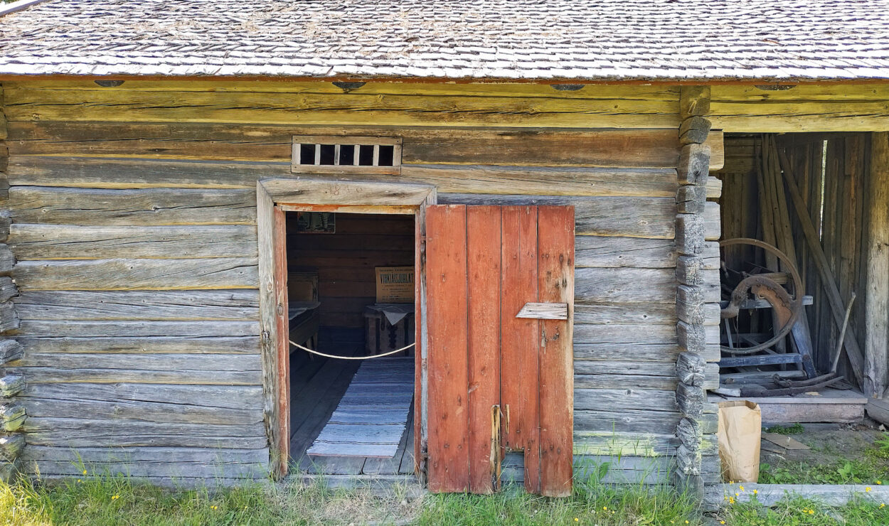 Eine verwitterte Holzhütte mit offener Tür und sichtbarer Eingangsstufe. Rechts ist ein Holzrad zu sehen. Die Hütte ist von Gras und Bäumen umgeben.