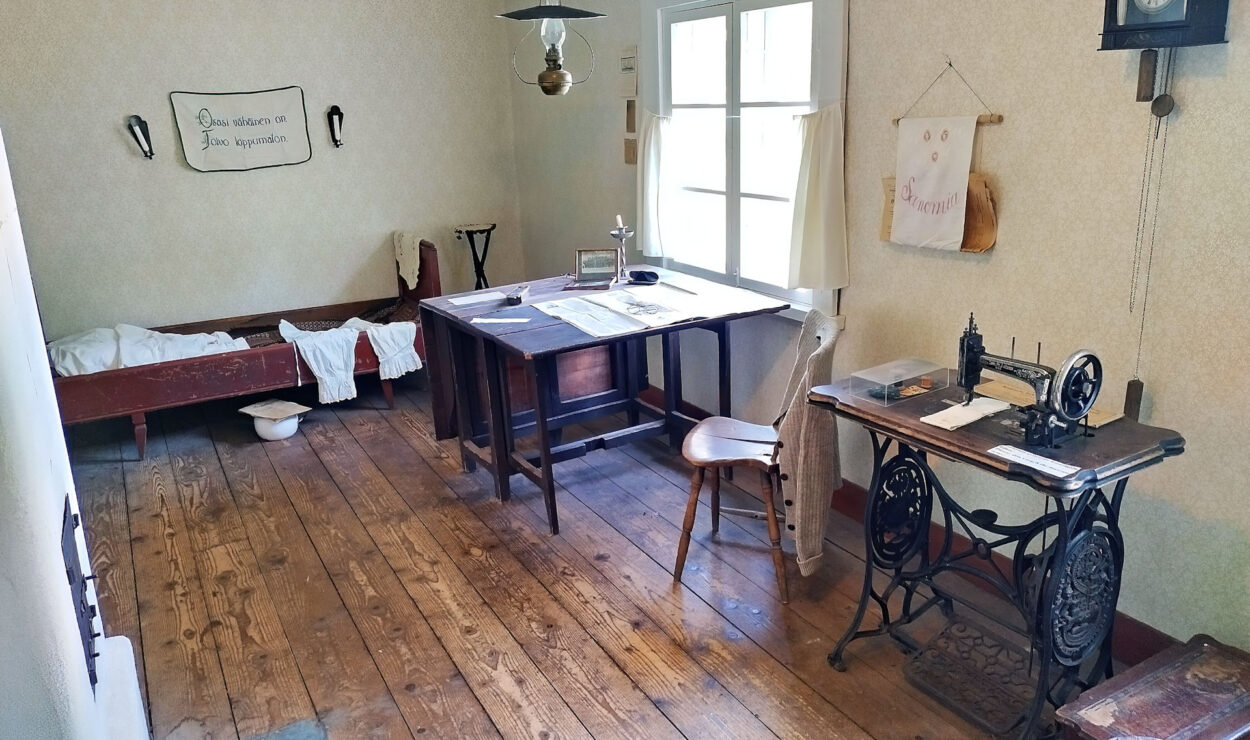 Vintage-Zimmer mit Holzfußboden, einem Nähmaschinentisch, einem Schreibtisch mit Papieren, einem Holzbett und einer Wanduhr. Es gibt natürliches Licht durch das Fenster und ein dekoratives Wandbehang.