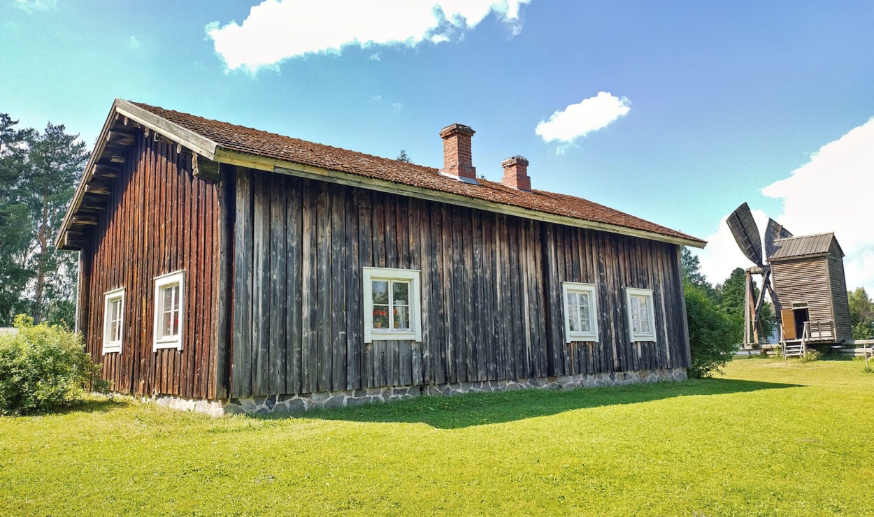 Ein rustikales Holzhaus mit rotem Dach steht auf einer Wiese unter blauem Himmel. Im Hintergrund ist eine Windmühle zu sehen.