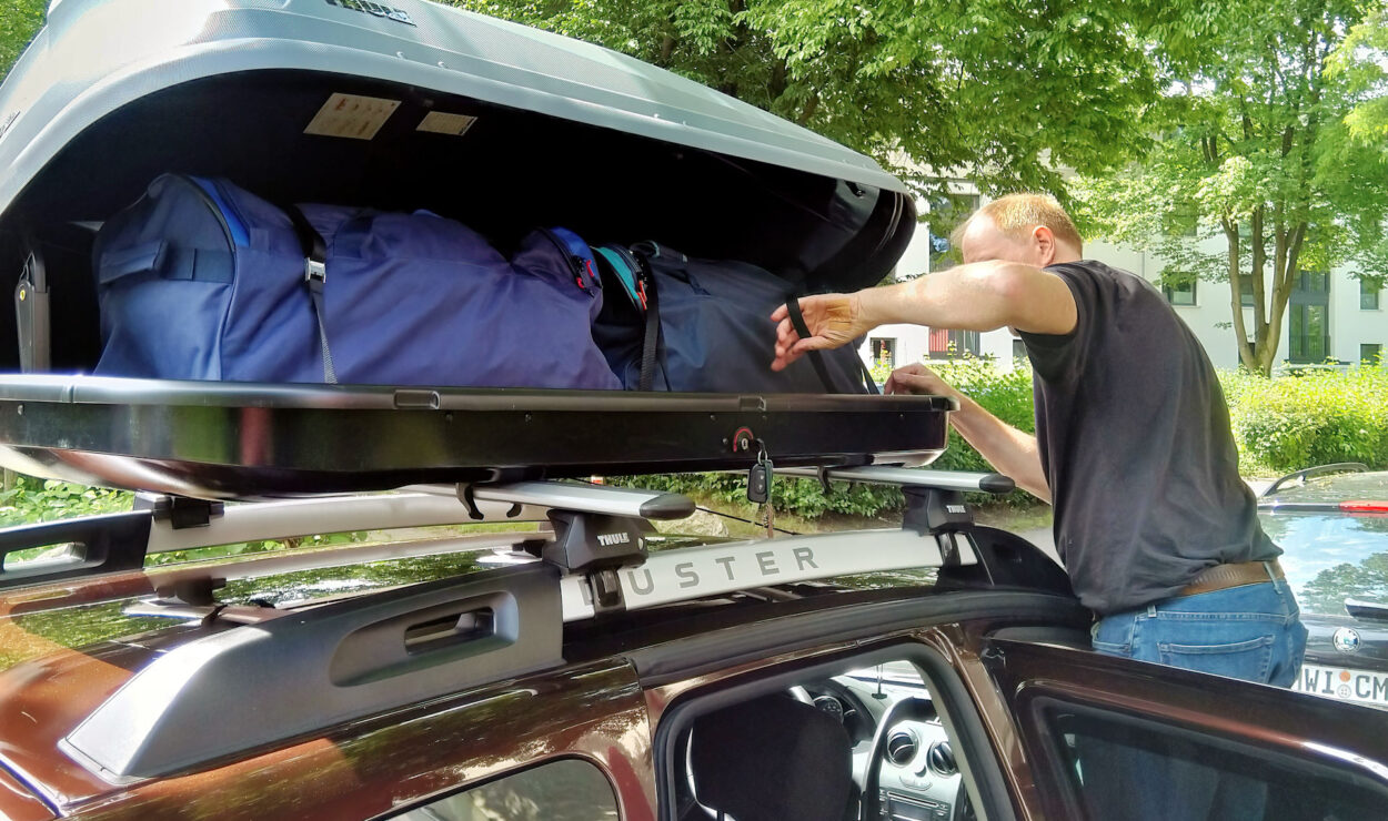 Ein Mann lädt Taschen in einen offenen Dachgepäckträger auf einem von Bäumen umgeben geparkten Auto.