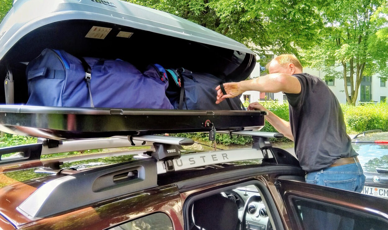 Person, die Taschen in eine Dachbox auf einem auf der Straße geparkten Fahrzeug legt, umgeben von grünen Bäumen.