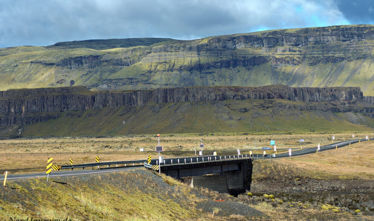 Eine Brücke überquert eine Straße in einer weiten, offenen Landschaft mit geschichteten Klippen in der Ferne unter einem bewölkten Himmel.