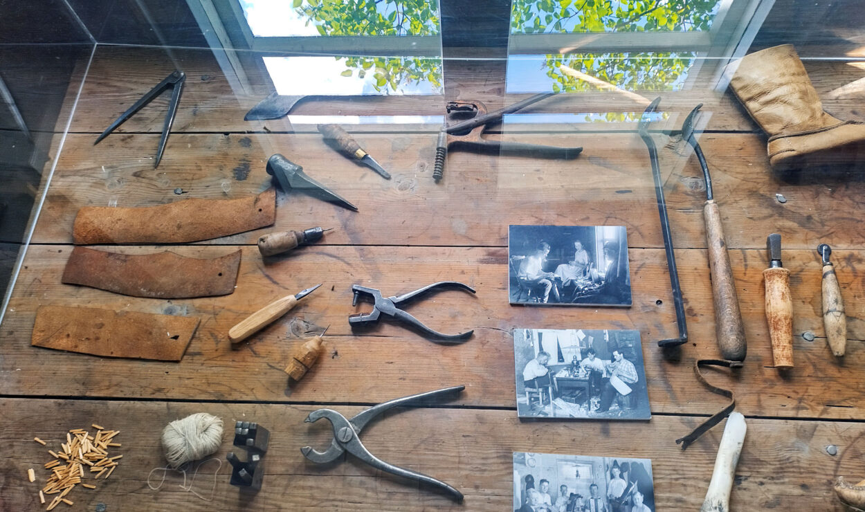 Eine Vitrine mit Vintage-Werkzeugen, Schwarzweißfotos und Holzobjekten auf einer Holzoberfläche. Im Glas spiegelt sich ein Blick aus dem Fenster ins Grüne.