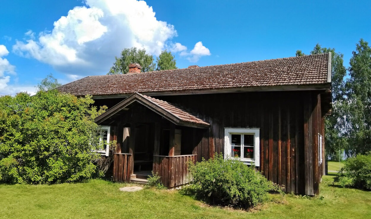 Eine rustikale Holzhütte mit Satteldach steht inmitten von grünen Büschen und Gras unter einem blauen Himmel mit Wolken.