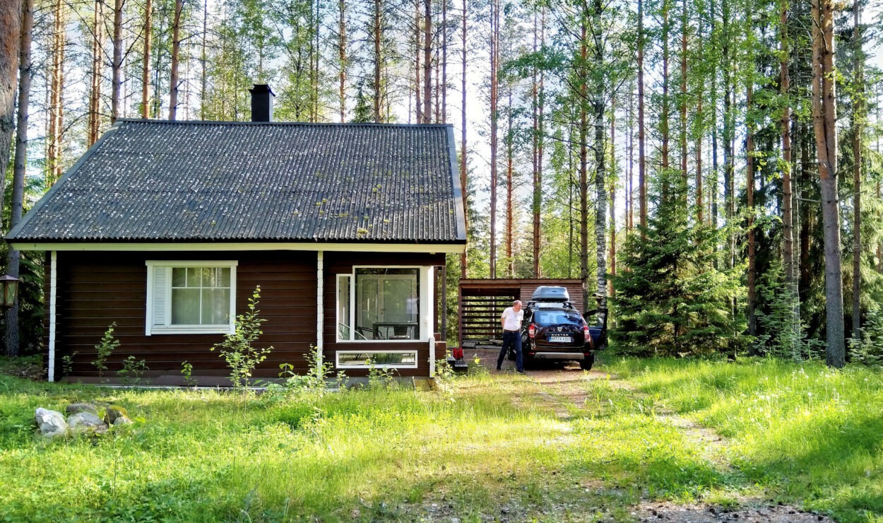 Eine kleine Holzhütte mit schrägem Dach, umgeben von hohen Bäumen. Neben der Hütte auf einer Rasenfläche parkt ein Auto, neben dem sich eine Person befindet.