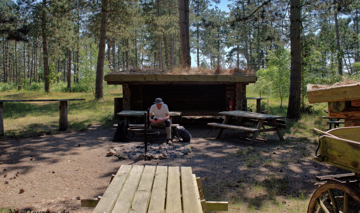 Person, die vor einem rustikalen Unterstand in einem Waldstück sitzt, umgeben von Picknicktischen und einem Holzkarren.