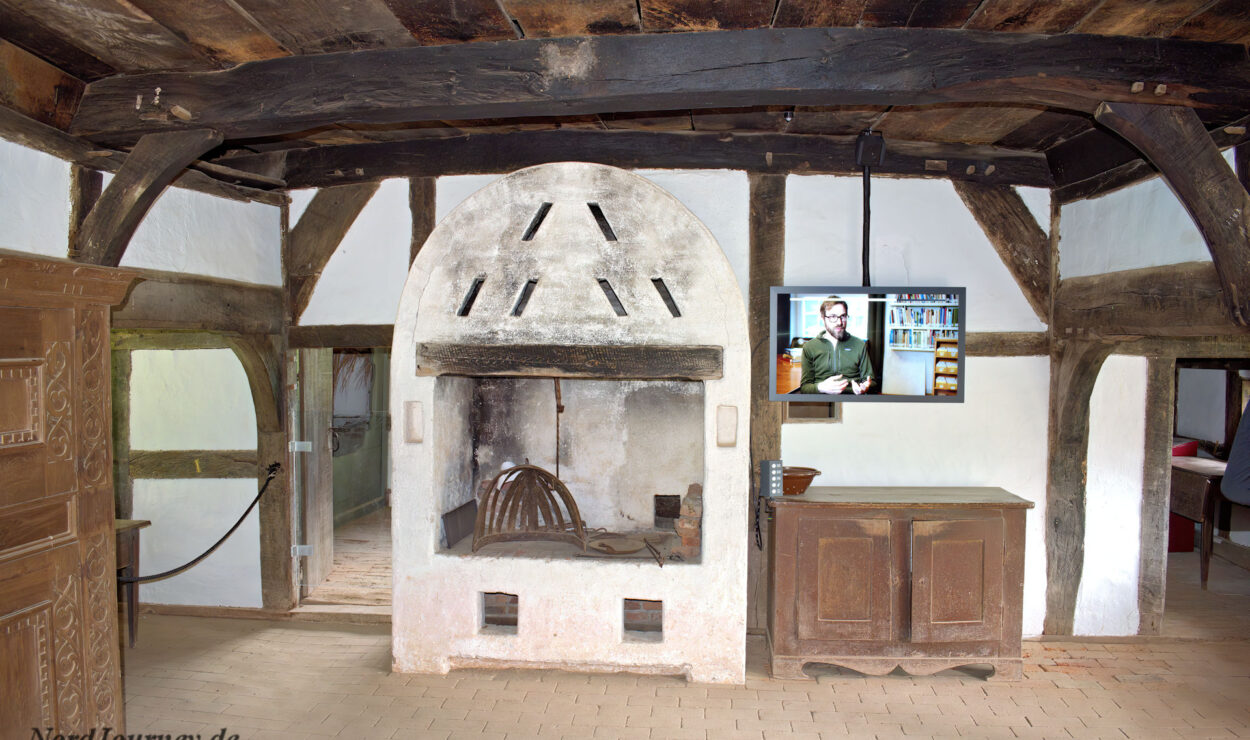 Ein rustikaler Raum mit einem großen alten Kamin, Holzbalken, einem Schrank und einem an der Wand montierten modernen Fernseher, auf dem eine Person und Regale zu sehen sind.