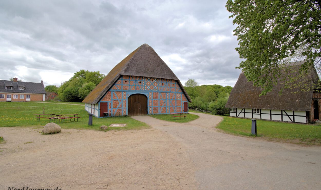 Eine traditionelle Fachwerkscheune mit Strohdach steht in ländlicher Umgebung im Freien, flankiert von Bäumen und weiteren Gebäuden unter einem bewölkten Himmel.