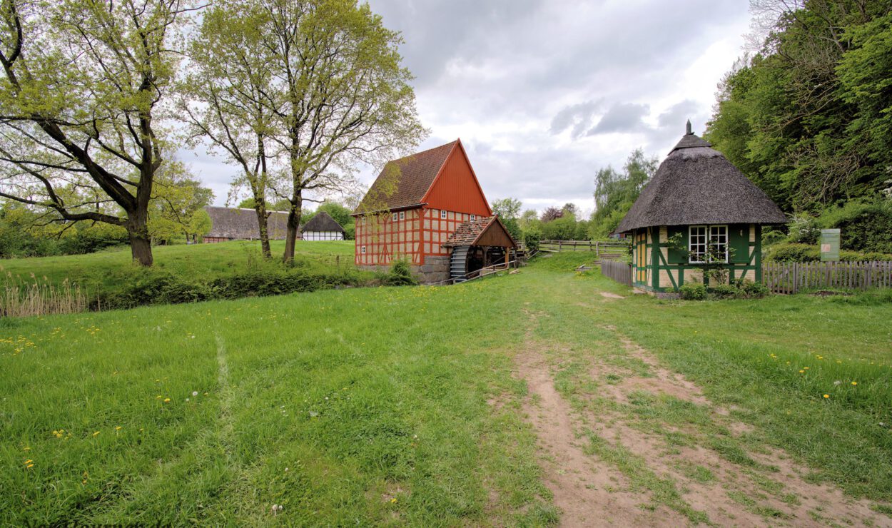 Eine Wiese mit einem Feldweg führt zu einem roten, scheunenartigen Fachwerkgebäude und einem kleinen grünen Haus mit Strohdach, umgeben von Bäumen und Holzzäunen.