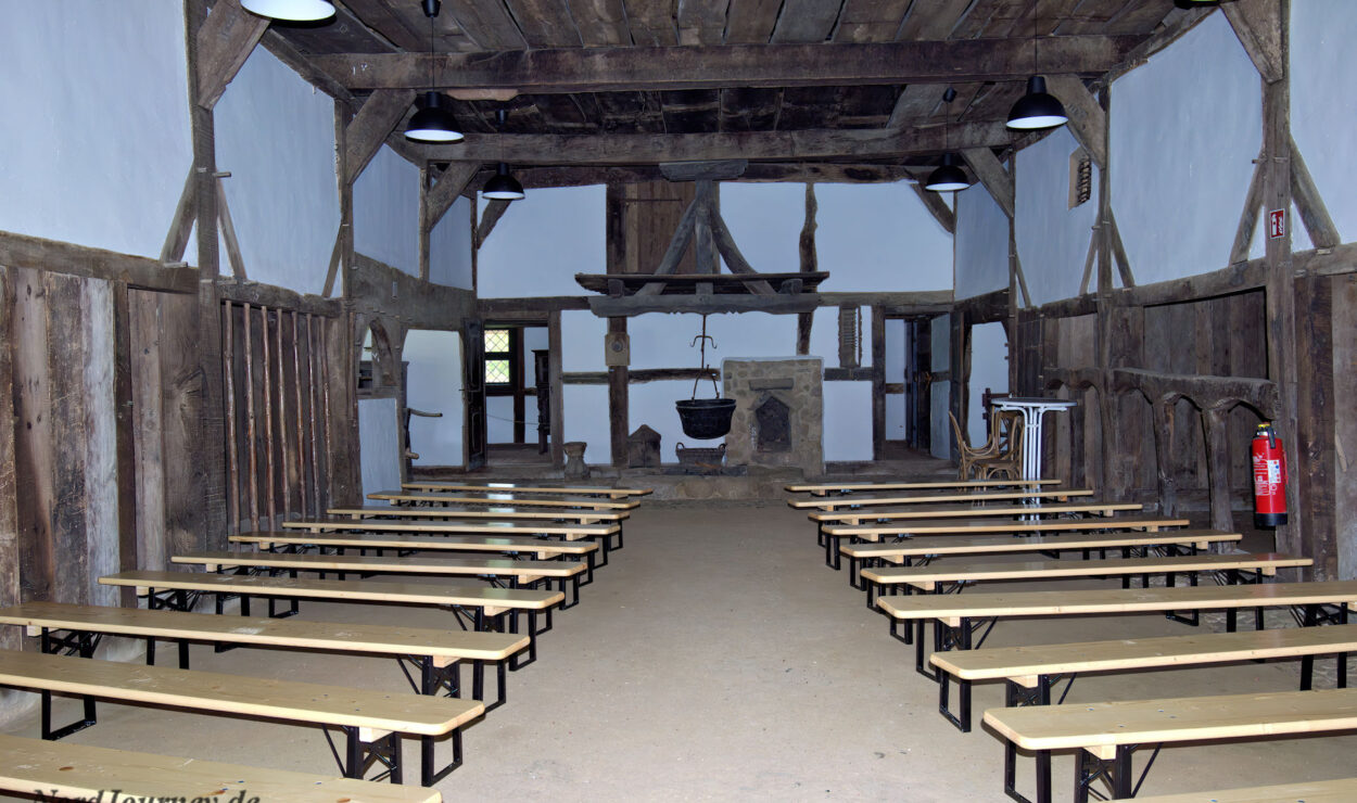 Ein rustikaler Saal mit Holzmöbeln und Reihen einfacher Bänke gegenüber einem Kamin und Herd am anderen Ende; freiliegende Balken und weiß getünchte Wände vervollständigen den Raum.