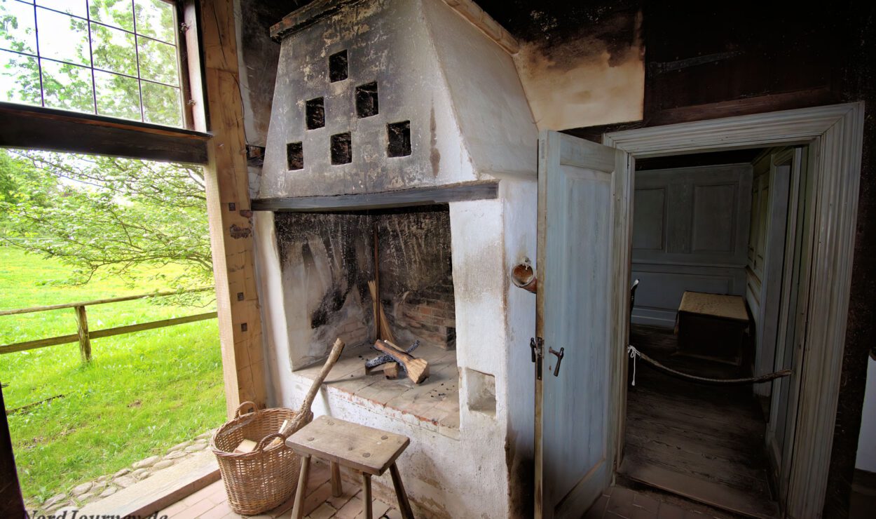 Altmodischer Innenkamin mit einem Korb Brennholz und einem kleinen Tisch daneben, neben einer offenen Tür, die in einen anderen Raum führt. Durch ein Fenster sieht man draußen grünes Gras und Bäume.