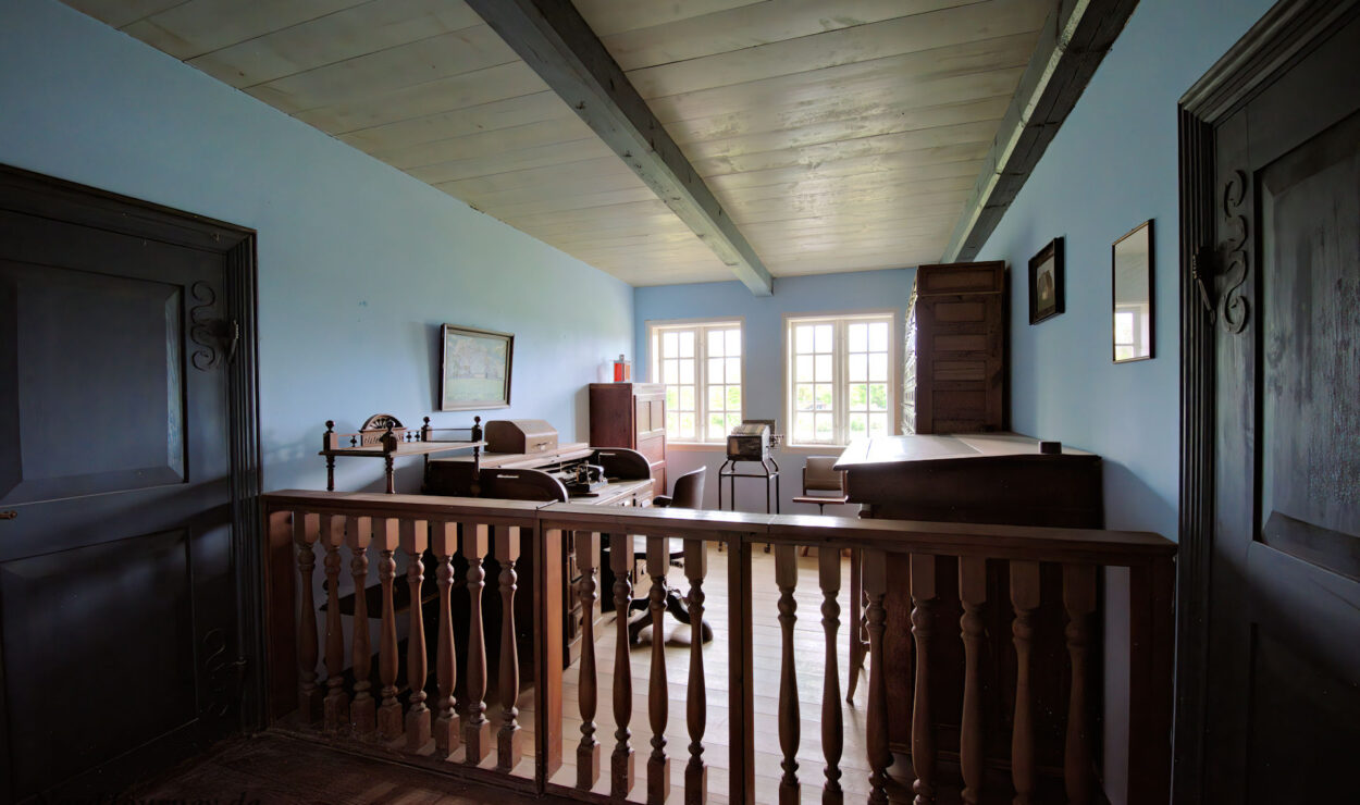 Ein Vintage-Büro mit Holzmöbeln, Schreibmaschine und großen Fenstern, die natürliches Licht hereinlassen; blaue Wände und ein Holzgeländer im Vordergrund.
