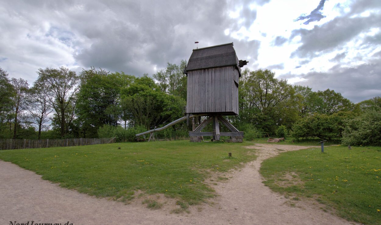 Eine hölzerne Windmühlenkonstruktion auf Stelzen in einem grasbewachsenen Park mit bewölktem Himmel, umgeben von Bäumen und Spazierwegen.