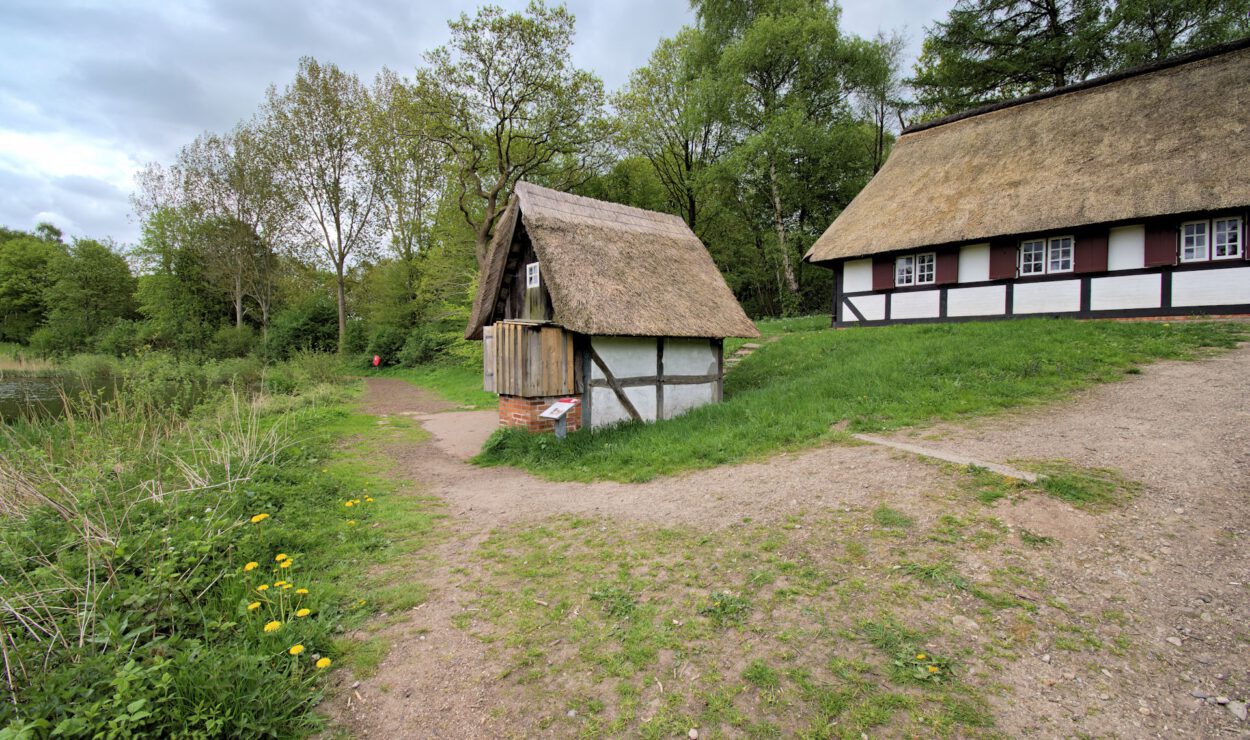 Ein kleines Reetdachhaus steht neben einem größeren Fachwerkhaus, umgeben von grünem Gras, Bäumen und einem Feldweg unter einem bewölkten Himmel.