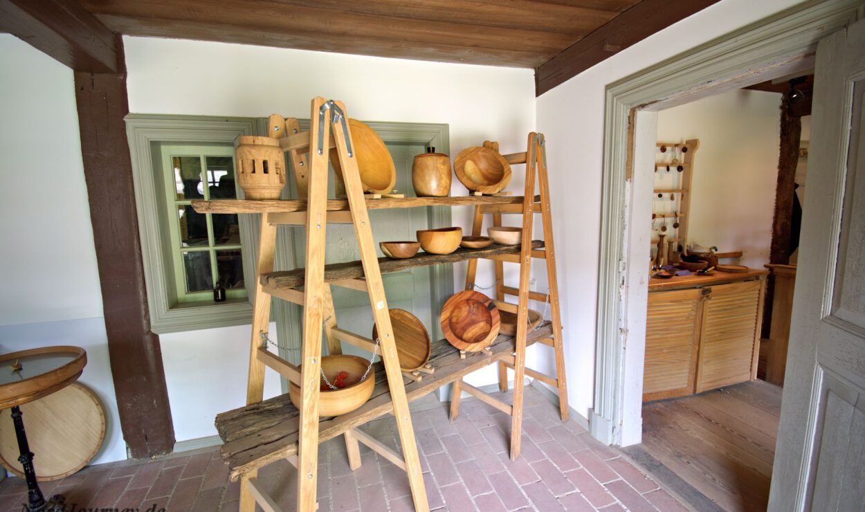 In einer Ecke stehen auf Holzregalen verschiedene Schüsseln und Utensilien aus Holz; eine Tür führt in einen anderen Raum, in dem ähnliche Holzgegenstände ausgestellt sind.