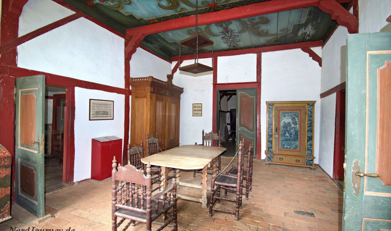 Historisches Esszimmer mit Holztisch und -stühlen, kunstvoll bemalter Decke, gefliestem Boden und dekorativem Kleiderschrank, durch eine offene Tür bei natürlichem Licht zu sehen.