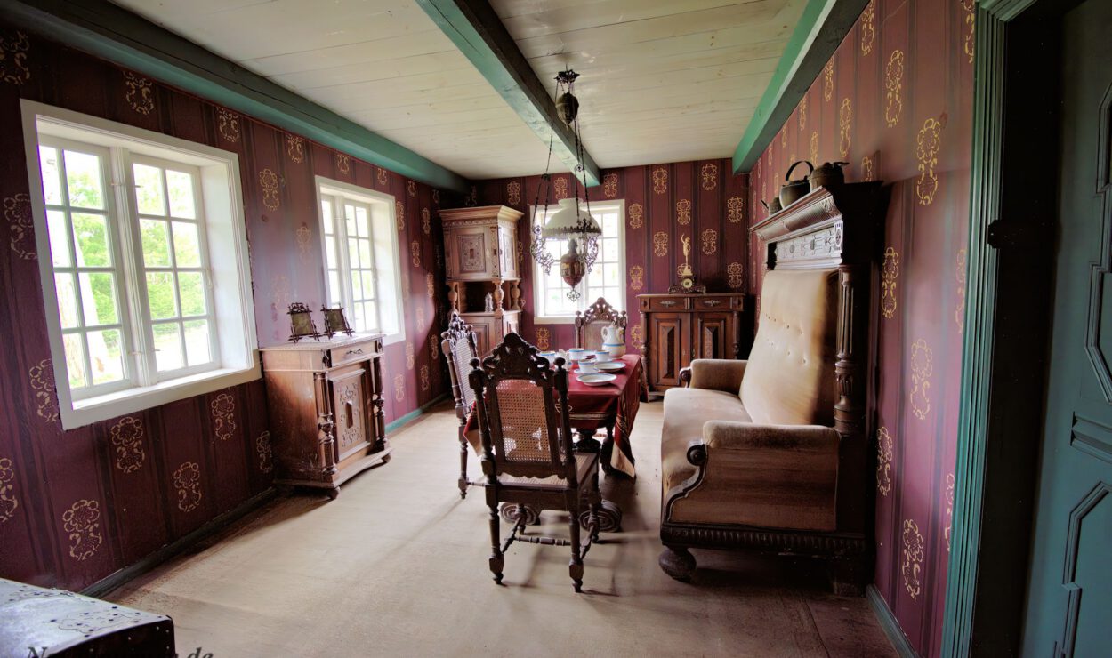 Ein Esszimmer im Vintage-Stil mit kunstvollen Holzmöbeln, gemusterten Tapeten und Sonnenlicht, das durch große Fenster strömt.