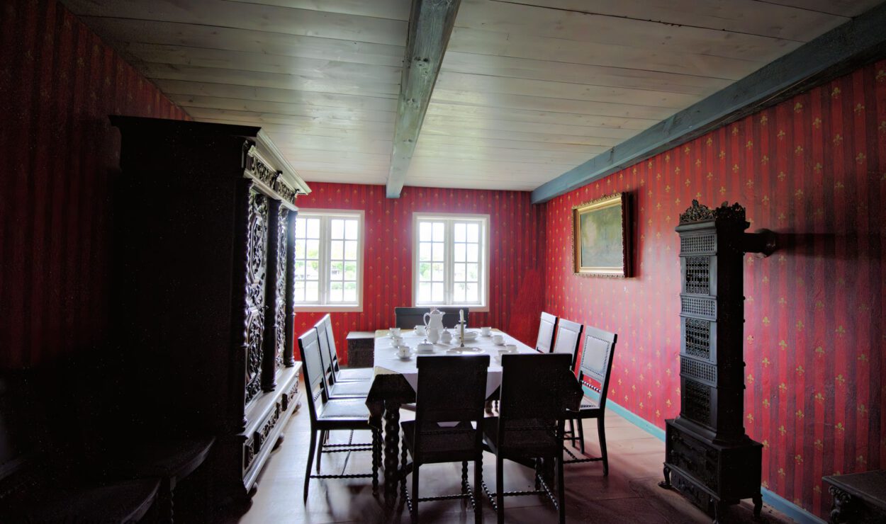 Ein Esszimmer mit einem langen Tisch, Stühlen, verzierten Holzmöbeln, gemusterter roter Tapete und Fenstern, die natürliches Licht hereinlassen.