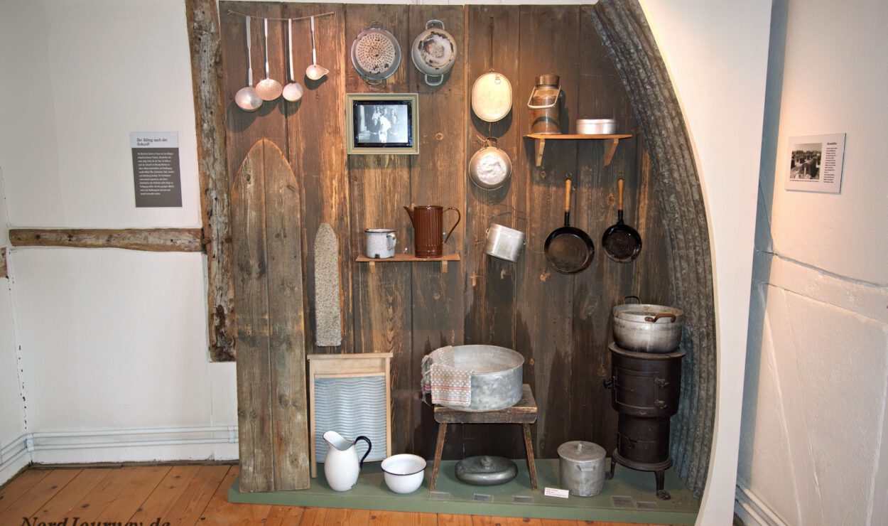 Eine Ausstellung von alten Küchenutensilien und Kochgeschirr, darunter Töpfe, Pfannen, Schöpfkellen und ein Waschbrett, an einer Holzwand in einem Museumsambiente.