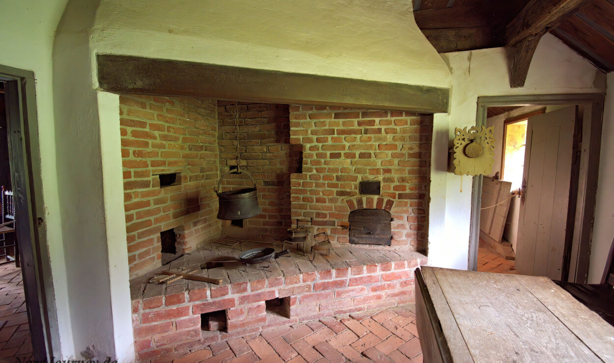 Historischer gemauerter Herd mit einem über dem Feuer hängenden Kessel, einer kleinen Ofentür und rustikalen Holzbalken in einer altmodischen Küche.