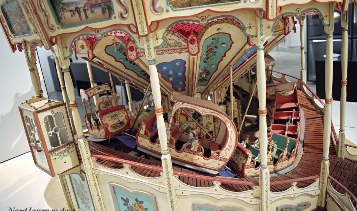 Detailliertes Modell eines klassischen Karussells mit kunstvollen Verzierungen, Miniaturfiguren und farbenfrohen gemalten Szenen auf den Paneelen.