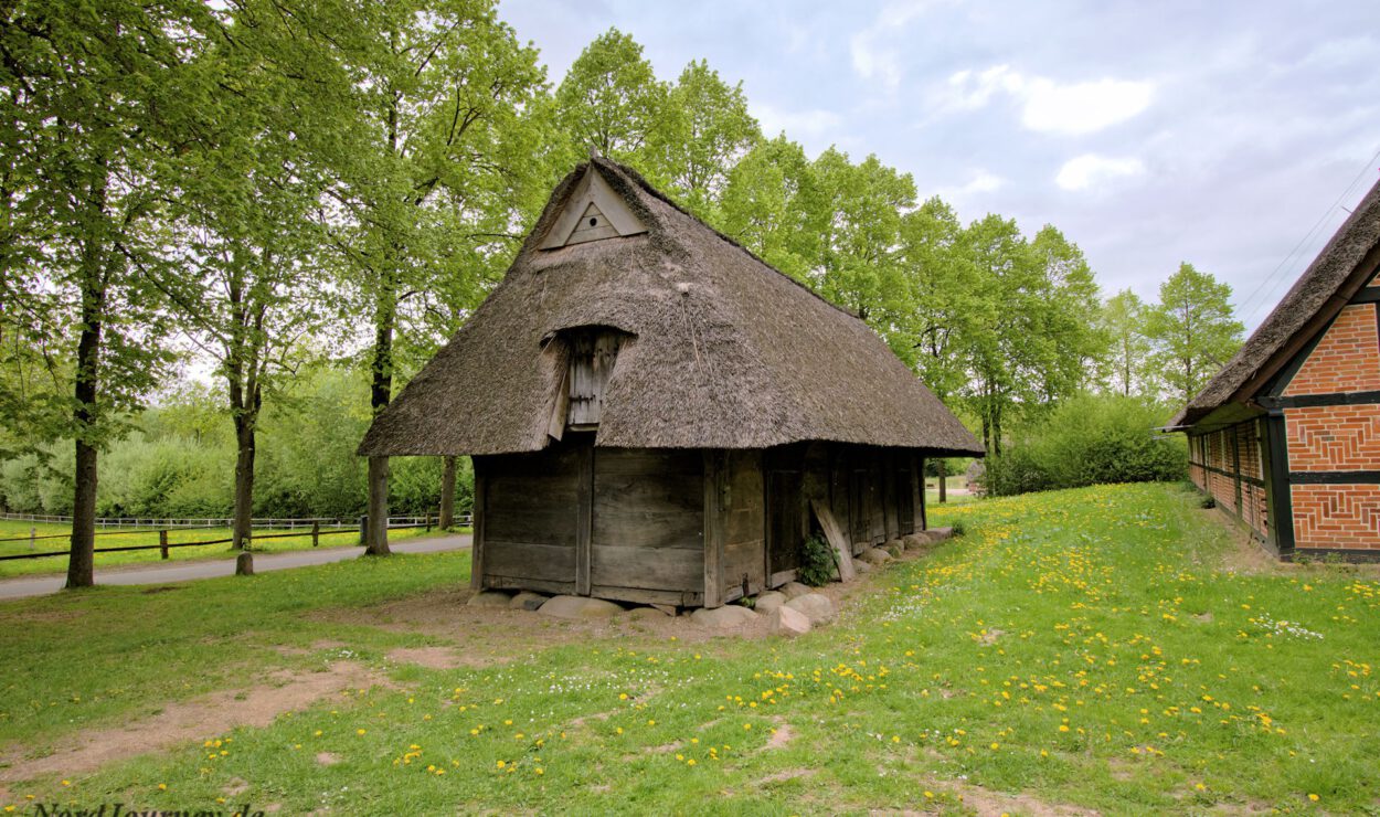 Auf einer Rasenfläche, umgeben von Bäumen, steht ein Holzgebäude mit steilem Strohdach. Rechts ist ein zweites Backsteingebäude teilweise sichtbar.
