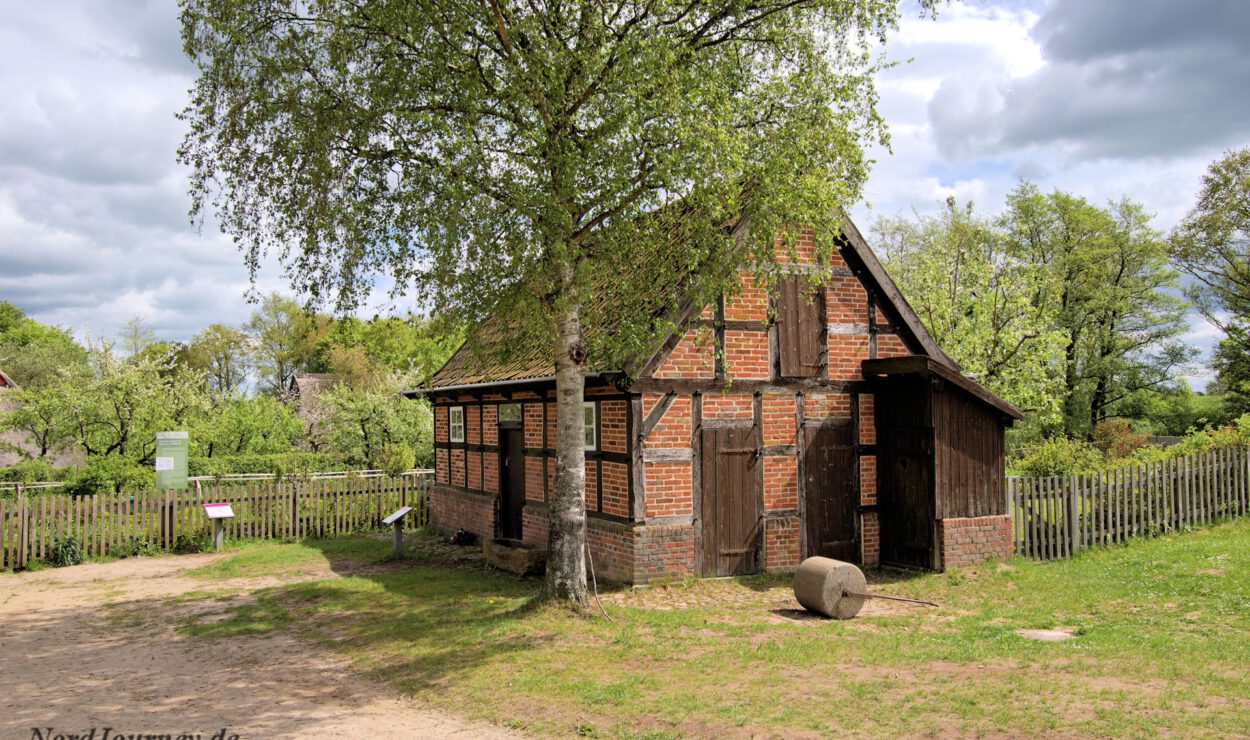 Ein kleines, altes Haus aus Backstein und Holz mit einem Holzzaun, einem Baum davor und einer grasbewachsenen Umgebung unter einem bewölkten Himmel.