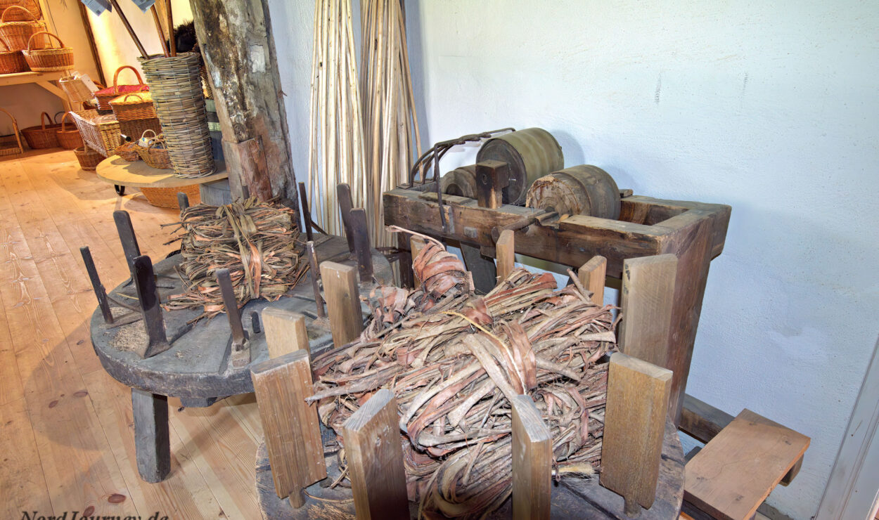 Eine Sammlung alter Holzwerkzeuge und Spindelgeräte zum Weben oder Korbflechten mit Bündeln von Rohstoffen in einer Werkstattumgebung.