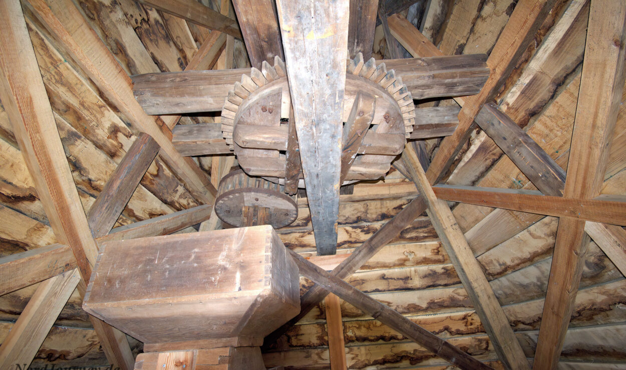 Innenansicht einer hölzernen Dachkonstruktion mit großen hölzernen Zahnrädern und Balken, möglicherweise Teil eines historischen Mühlenmechanismus.