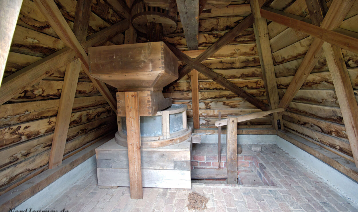Innenansicht einer hölzernen Windmühle mit freiliegenden Balken, Zahnrädern und Mühlsteinmechanismus auf einem Ziegelboden.