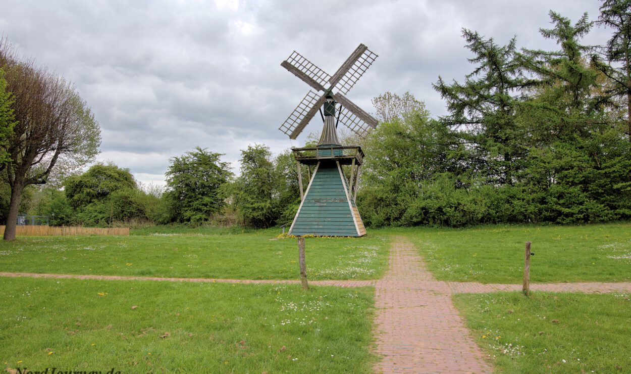 Eine traditionelle Windmühle aus Holz steht auf einer Wiese, zu der ein Ziegelweg führt; im Hintergrund sind Bäume und ein bewölkter Himmel zu sehen.