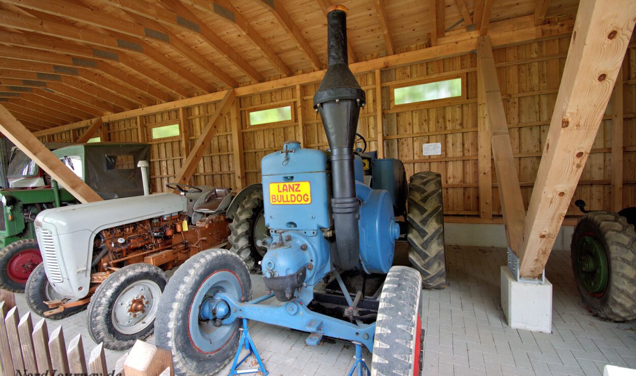 In einem Holzschuppen mit Satteldach sind ein blauer Lanz Bulldog Traktor und ein älterer grauer Traktor ausgestellt.