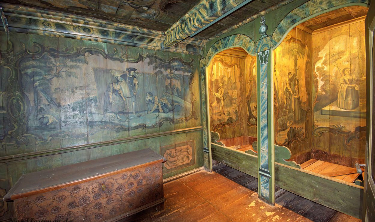 Reich verzierter, historischer Holzraum mit Wand- und Deckenmalereien, zwei offenen Toilettensitzen aus Holz und einer dekorativen Holztruhe.