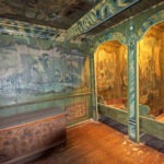 Reich verzierter, historischer Holzraum mit Wand- und Deckenmalereien, zwei offenen Toilettensitzen aus Holz und einer dekorativen Holztruhe.