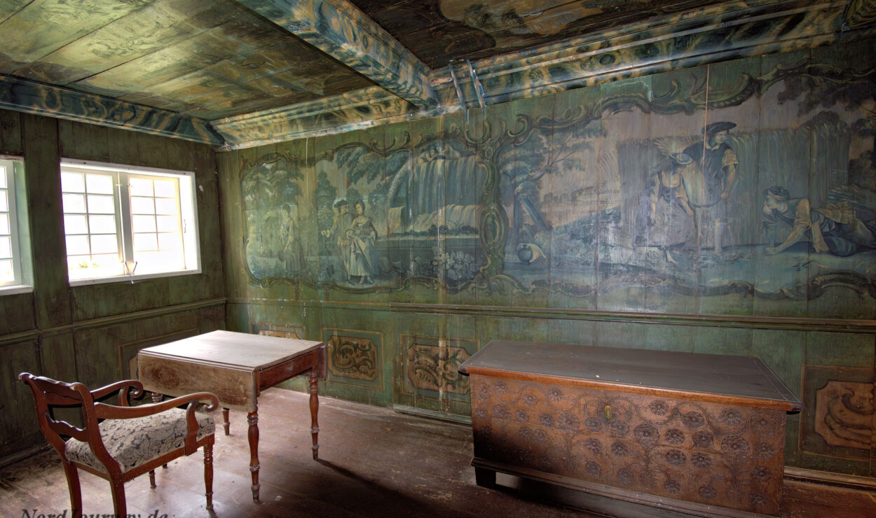 Ein historischer Raum mit grün gestrichenen Wänden und Decke, mit einem Holzstuhl, zwei Holztischen und einer großen Truhe unter einem Fenster.