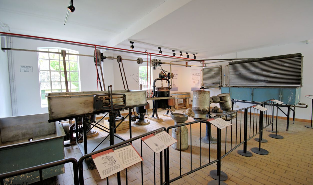 Eine historische Innenausstellung zeigt alte Industriemaschinen und -geräte hinter Geländern, mit Informationsschildern und einem Fenster, das natürliches Licht hereinlässt.