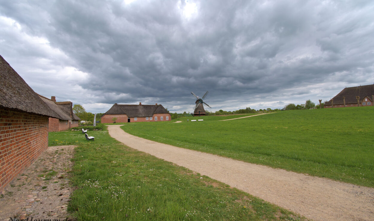 Ein Schotterweg führt durch eine Grasfläche zu einer Windmühle, umgeben von Backsteingebäuden unter einem bewölkten Himmel.