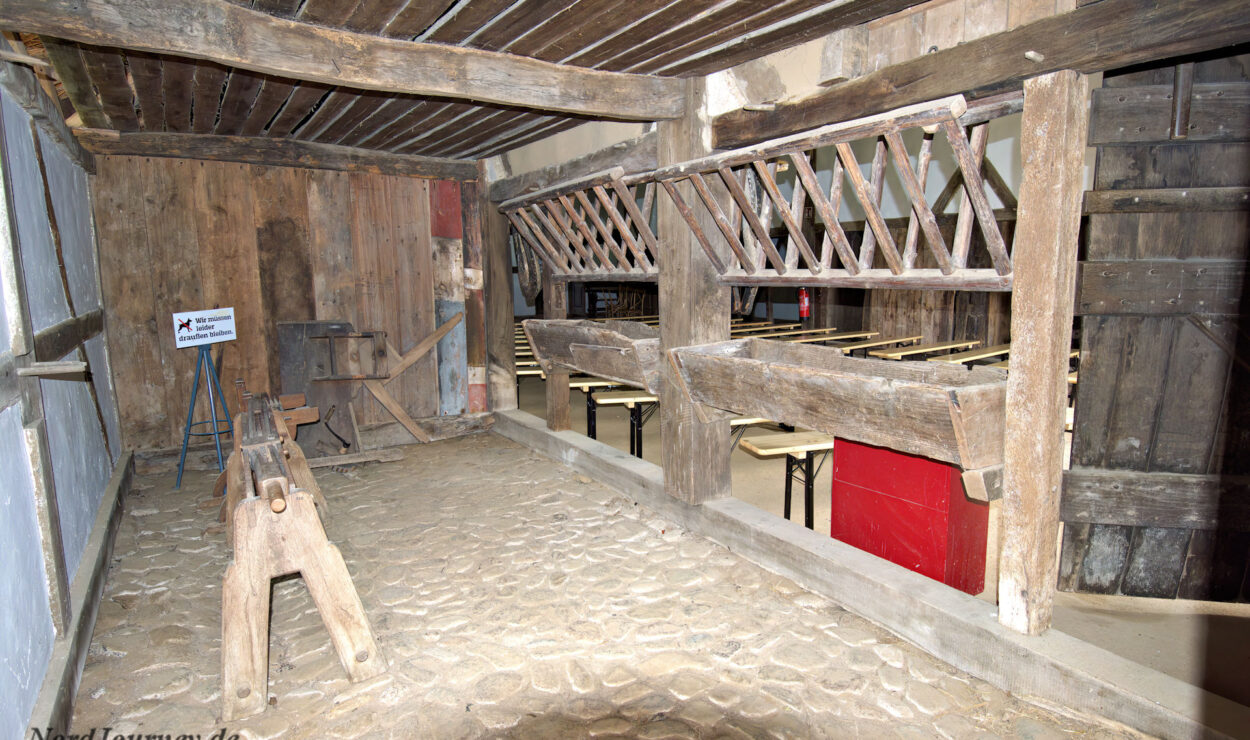 Innenansicht eines alten Stalles mit Holzboxen, Kopfsteinpflasterboden und Futterraufen. Im Hintergrund sind ein Schild und einige Bänke zu sehen.