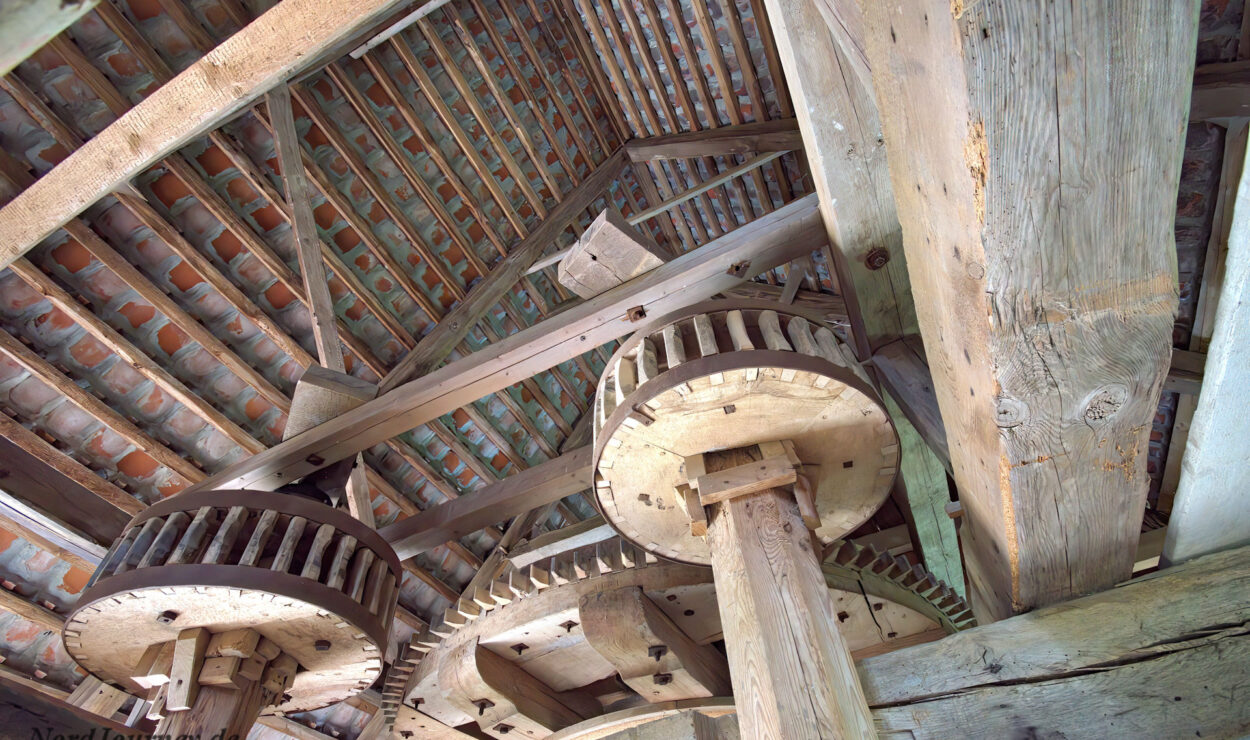 Hölzerne Zahnräder und Balken bilden den inneren Mechanismus einer traditionellen Windmühle, der unter einem Ziegeldach sichtbar ist.