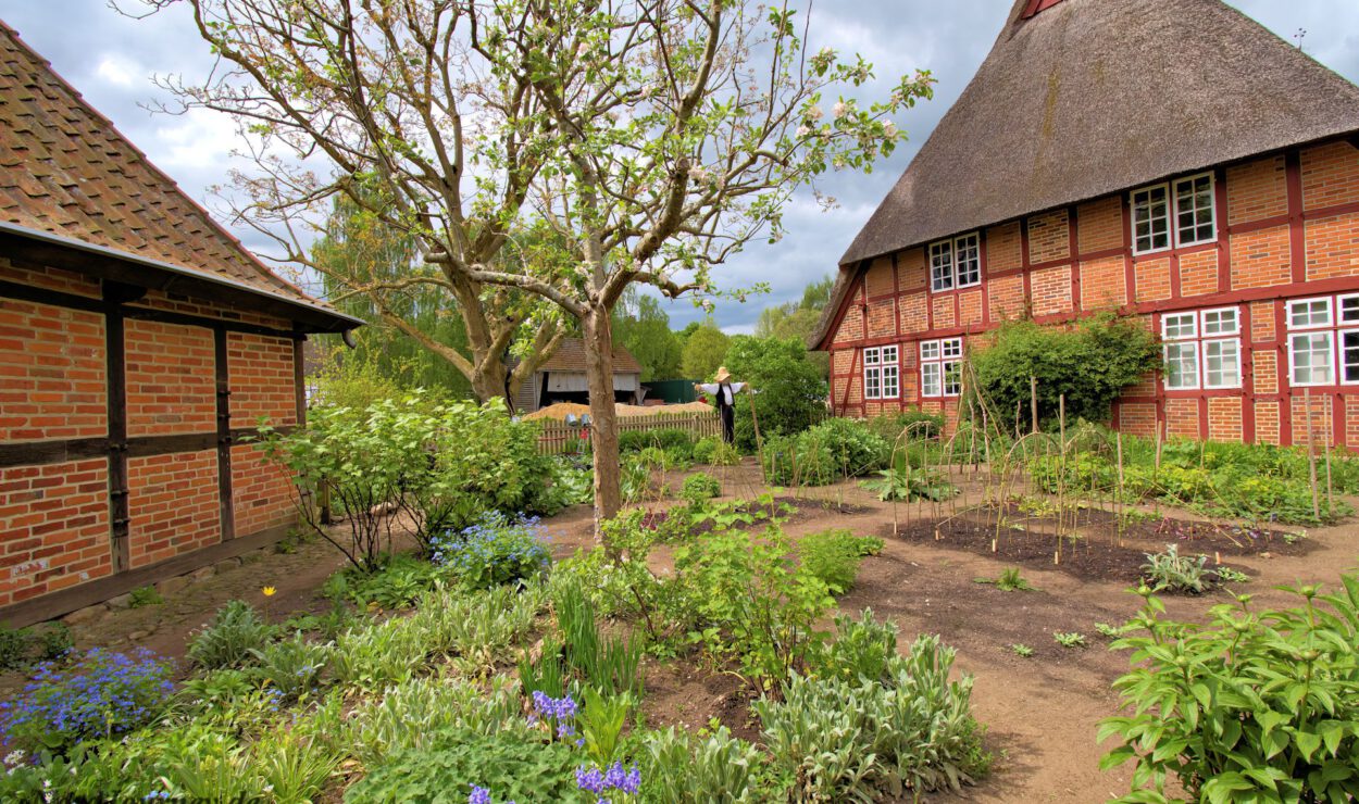 Ein traditionelles Haus aus Ziegeln und Holz mit Strohdach steht neben einem üppigen Garten mit verschiedenen Pflanzen, einem Baum und Blumenbeeten unter einem teilweise bewölkten Himmel.