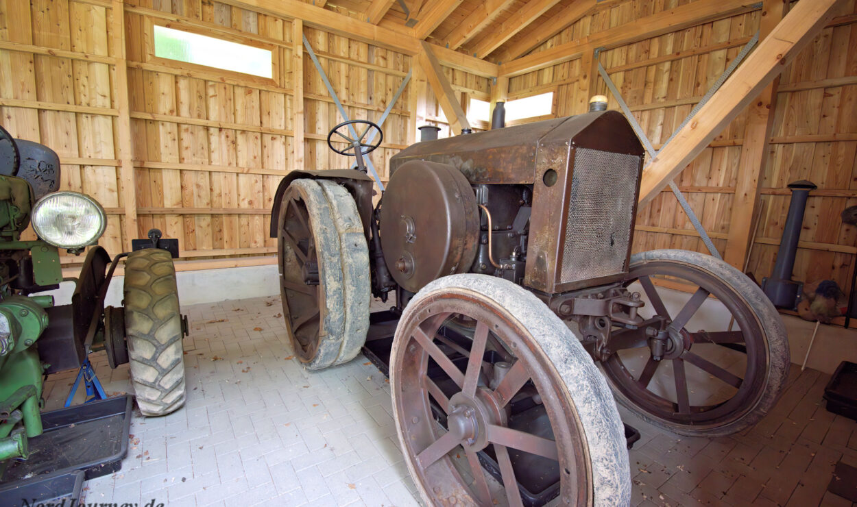 Ein alter, verrosteter Traktor mit Metallrädern steht in einem Holzschuppen mit Fliesenboden. Links ist ein weiterer grüner Oldtimer-Traktor teilweise sichtbar.